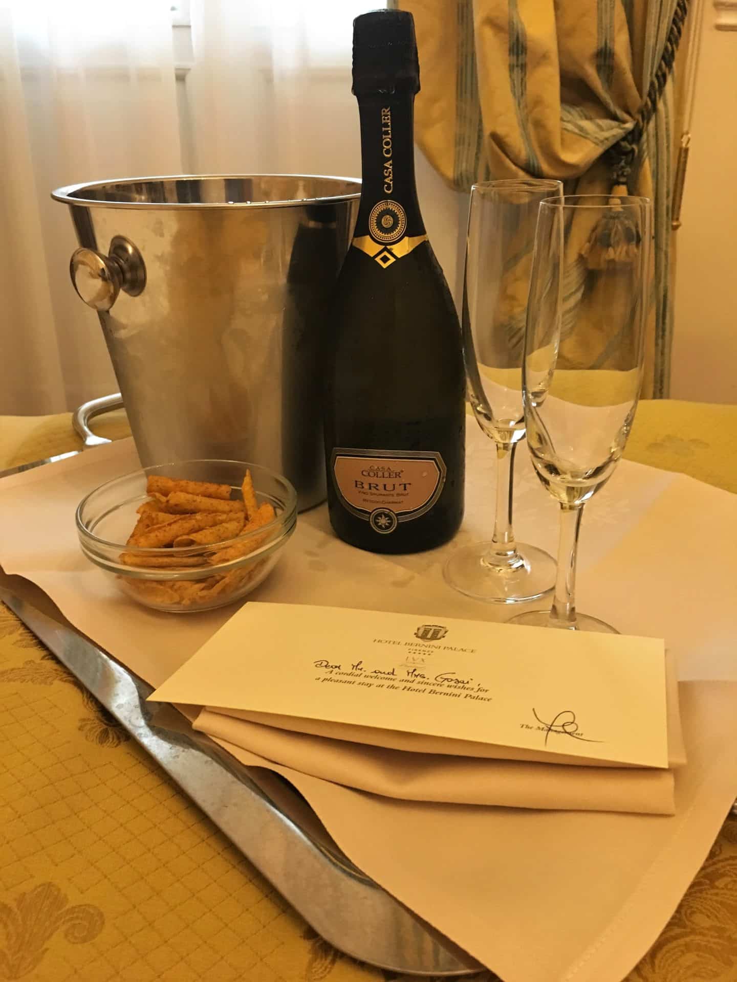 The Opulent Bernini Palace Hotel suoerior bedroom with some hotel stationery, a bottle of Champagne and 2 champgagne flutes.