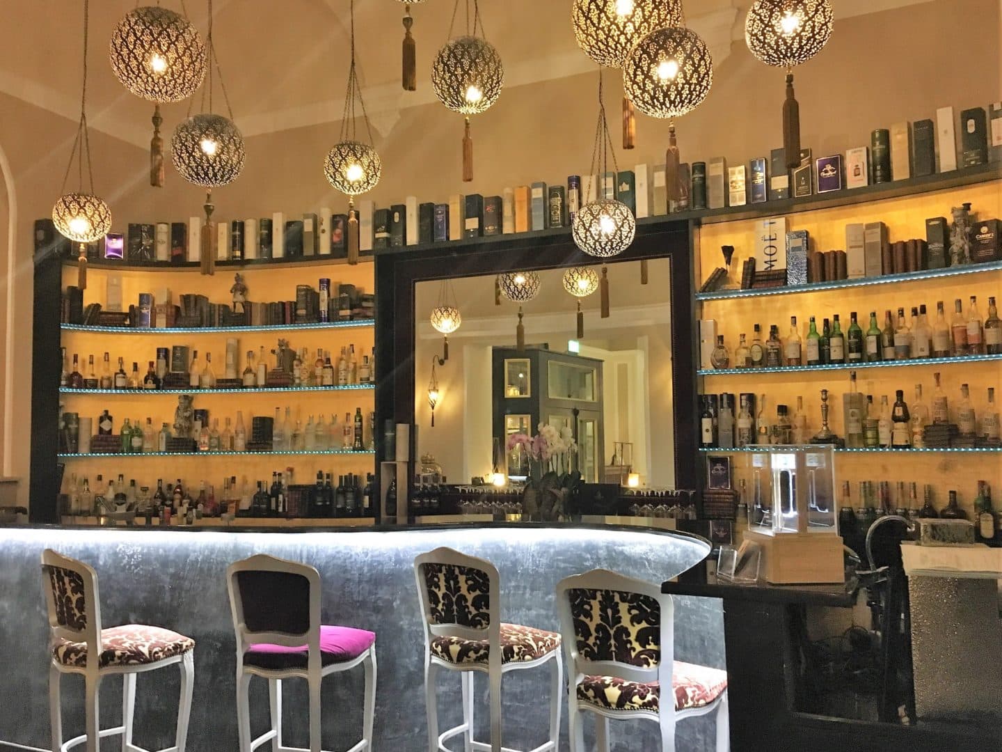 The beautiful Bernini Palace Hotel bar with magenta and gold seats arranged around a white topped bar table. Drinks are on shelves on a unit behind against a yellow wall. There are circular antique gold hanging down from the ceiling. The hotel is located in the heart of Florence