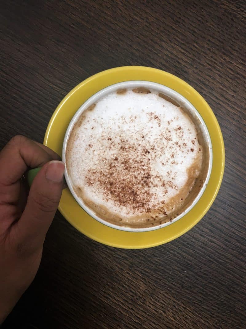 LE1:Independent Coffee & Cake Locales. Bru Coffee and Gelato. A cappuccino in a yellow cup with Bejal's hand on the handle, Leicester