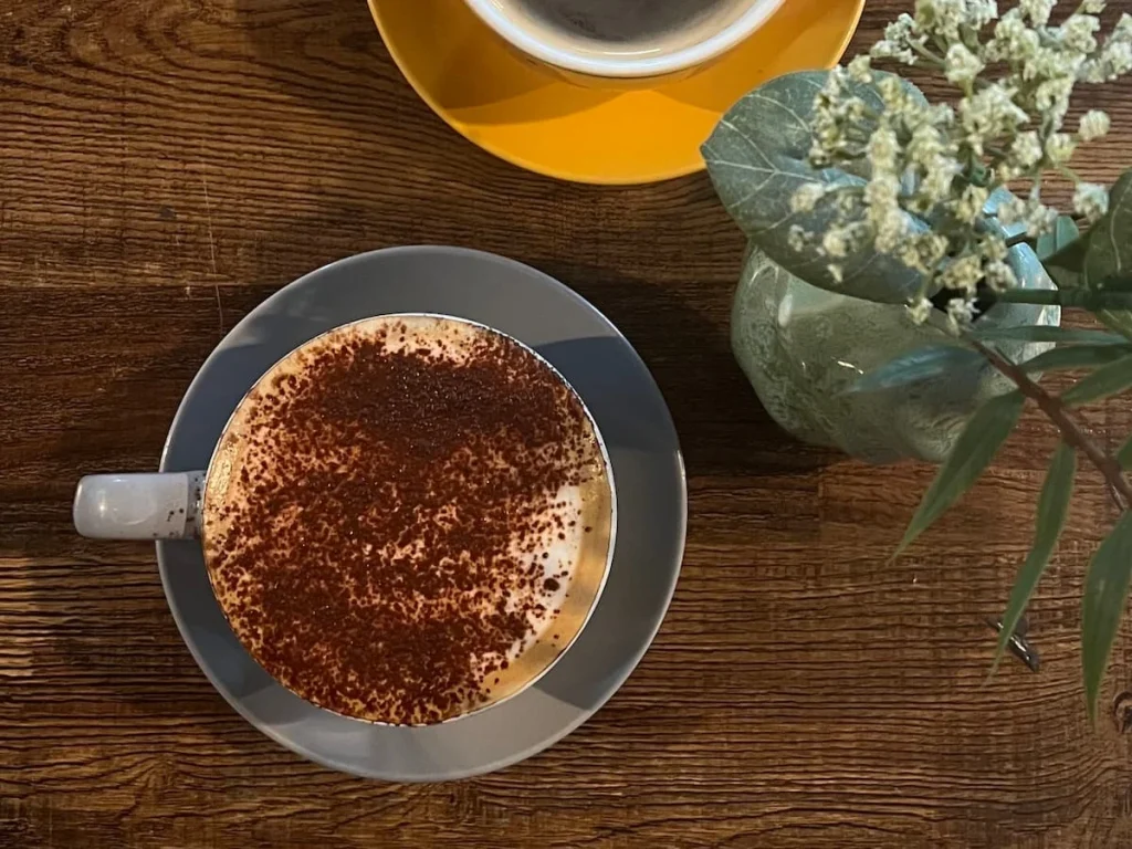 Black coffee and cappuccino flat lay at Lynn, Leicester. The image is a flat ay of a yellow coffee cup and saucer with a black coffee and a grey coffee cup and saucer with a cappuccino in it on a dark brown table top. There are some small jib flowers in a grey vase at the side