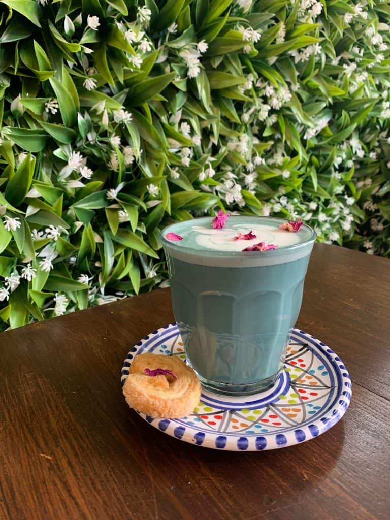 Blue coloured Luna Latte at la Foret Cafe with red rose petal decoration. The latte is begind a leaf and flower wall green background
