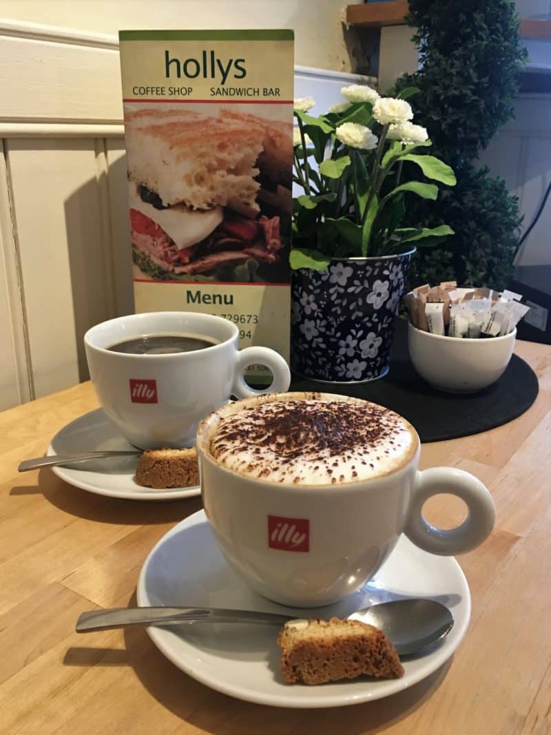 LE1:Coffee & Cake Locales. Hollys Coffee Shop & Sandwich Bar. 1 cappuccino and 1 black coffee with amoretto biscuits and a Holly's menu in the background.
