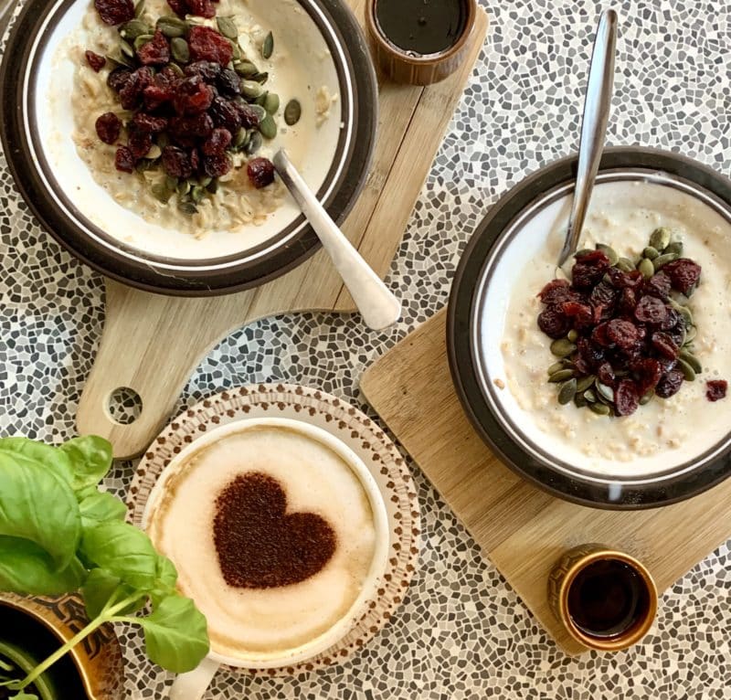 Independent Coffee Shops in LE1, Leicester. 2 x Porridge bowls topped with fruit and a pot of syrup, There is a cappuccino topped with a stencil chocolate heart. The items are on a speckled black and white background at Bryter Moon Deli