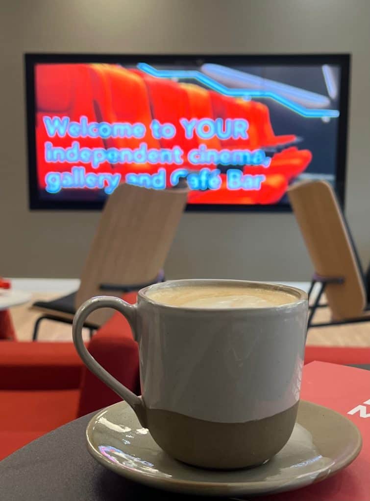 Latte in a mug with the red background on a TV screen reading, 'Welcoem to your Independent cinema, gallery and cafe bar' at The Phoenix, Leicester.