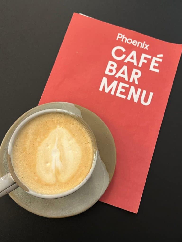 A Flat lay of a latte with the Phoenix cafe Bar Menu in red on a black table at Phoenix Arts Cafe, Leicester.