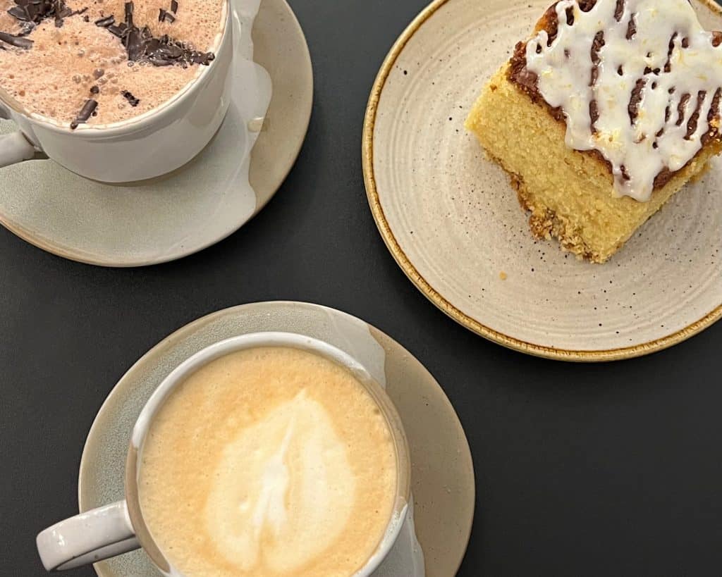 A flat lay of a latte, hot chocolate and lemon drizzle cake on a black background at The Phoenix Arts Cafe, Leicester