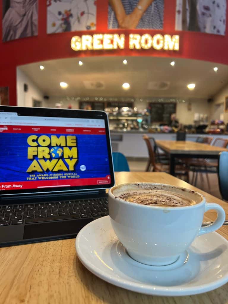 Cappuccino at the The Green Room, Curve, Leicester. There is an ipad in the image with the show text 'Come from Away' on the screen as well as the counter of the Green Room cafe and the signage lit up in bright light bulbs.
