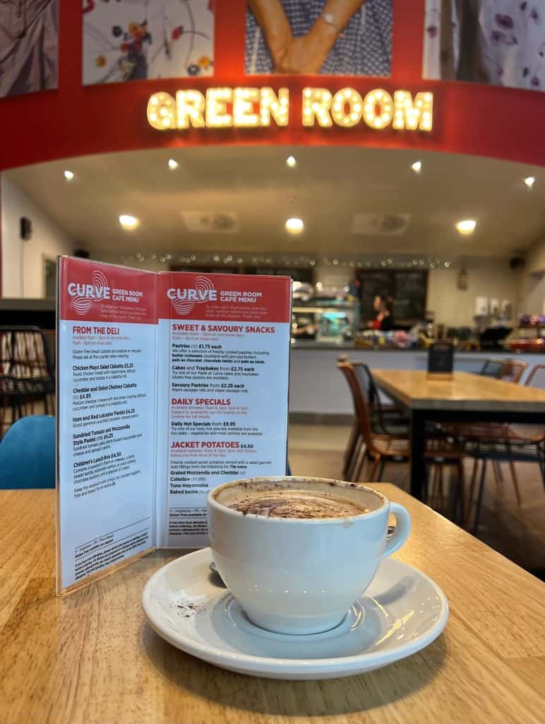 Cappuccino at the The Green Room, Curve, Leicester. There is a food menu in the image. The Green Room cafe is in the background and the signage lit up in bright light bulbs can be seen.