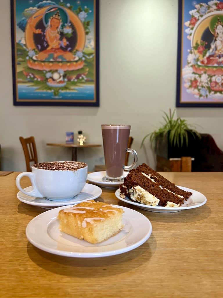 Lemon drizzle cake, hot chocolate, gluten free carrot cake on a table in the World Peace Cafe with a meditation photo on the wall.