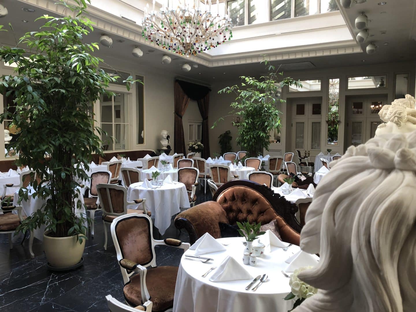 The Grand Palace Hotel Riga Orangery breakfast room filled with white tablecloth covered tables and red ornate chairs and chaise longue. Plants and porcelain cherubs are placed all around the room as well as a big chandelier in the centre.