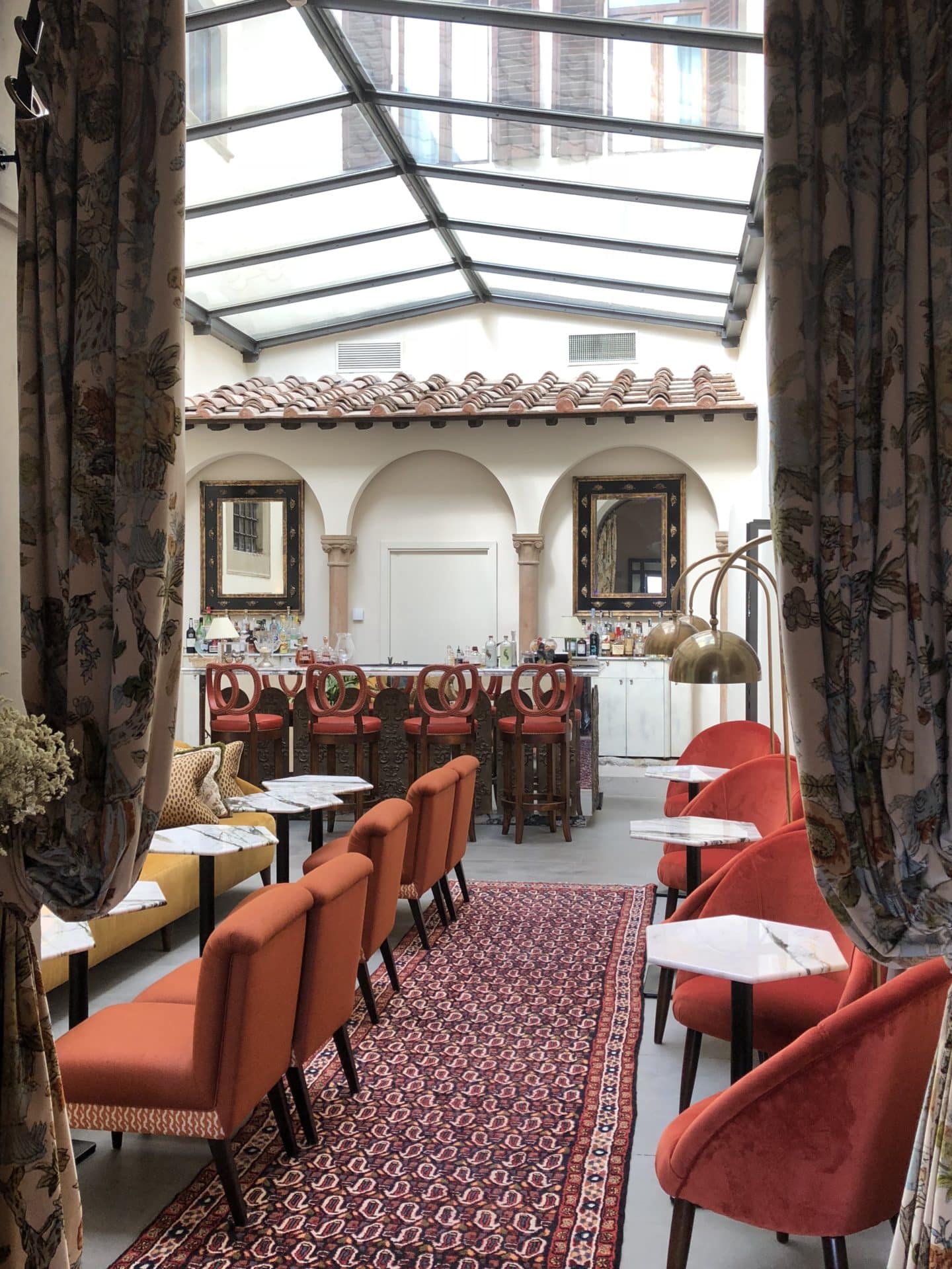 The archway into bar area lined with terracotta chairs, draped brown curtains and dark wood side boards. the bar can be seen at the top with bar stools surrounding it
