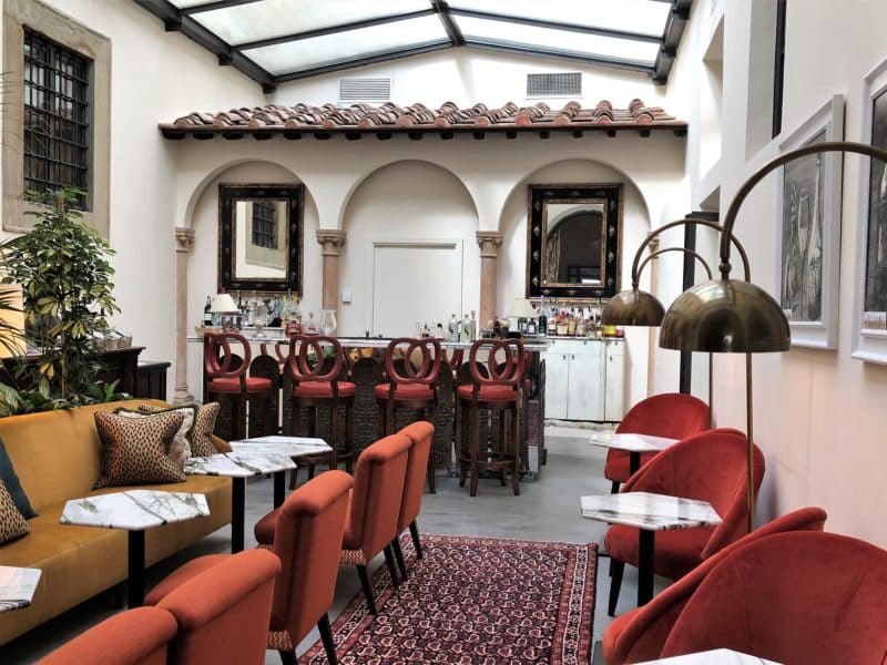The bar area lined with terracotta stools, adn arch lights as well as mustard sofas, marble topped circular tables and a corrugated roof. There are a few plants to the left of the bar area.