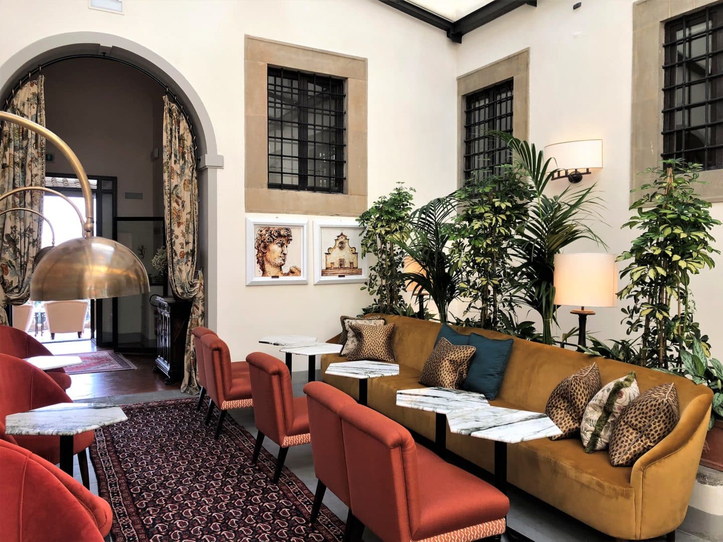 Brown and teal cushiosn on the sofe behind marble topped bar tables with tall plants in the background. The bar room at Belmond Villa San Michele.