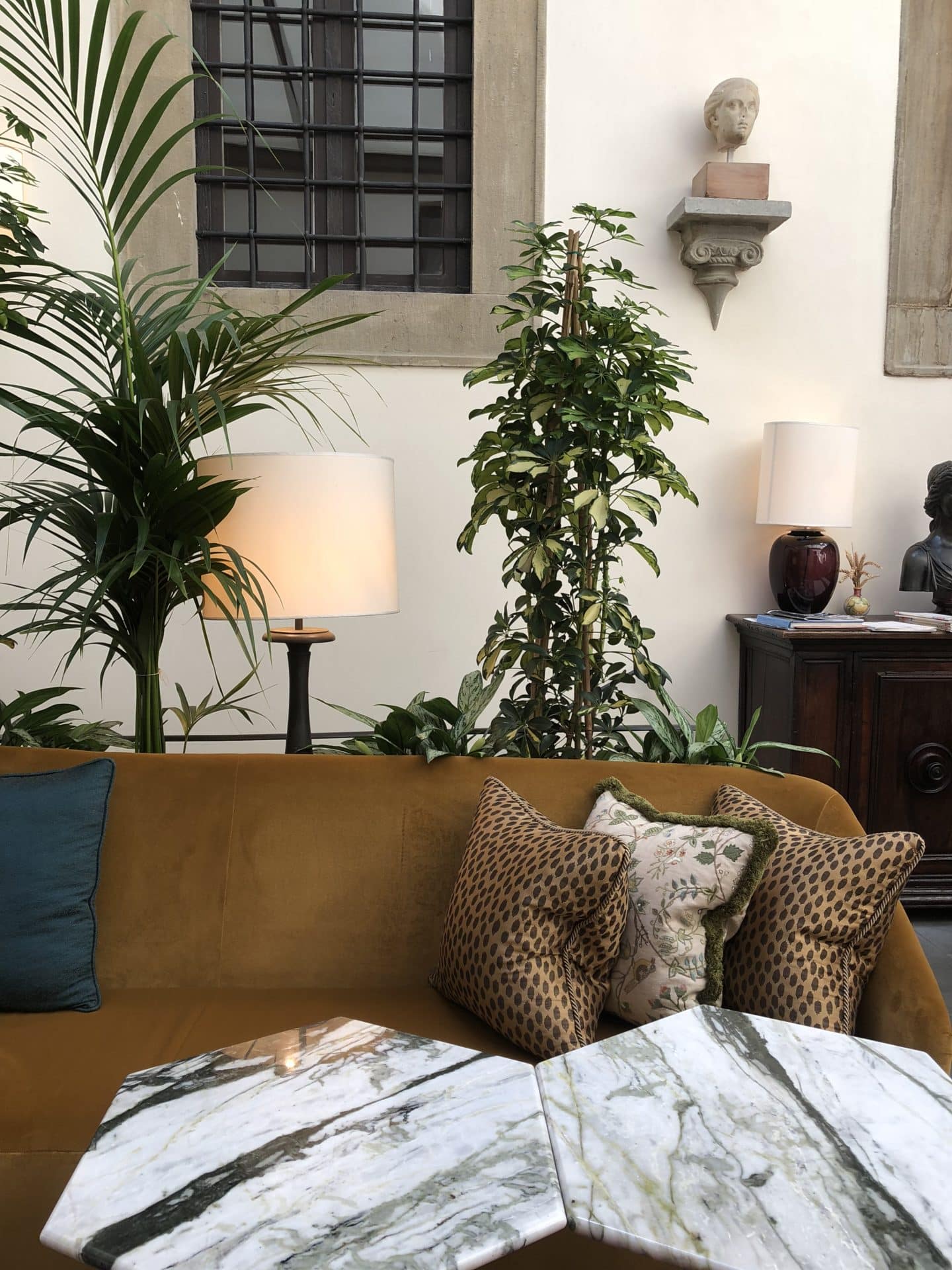 Brown and teal cushiosn on the sofe behind marble topped bar tables with tall plants in the background. The bar room at Belmond Villa San Michele.