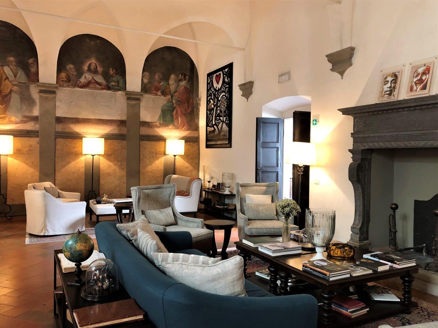 The lounge area at Belmond Villa San Michele wuth comfy sofas and armchairs covered in cream and blue linen fabric. Frescos on the upper walls with a dark green fireplace in the centre of the room