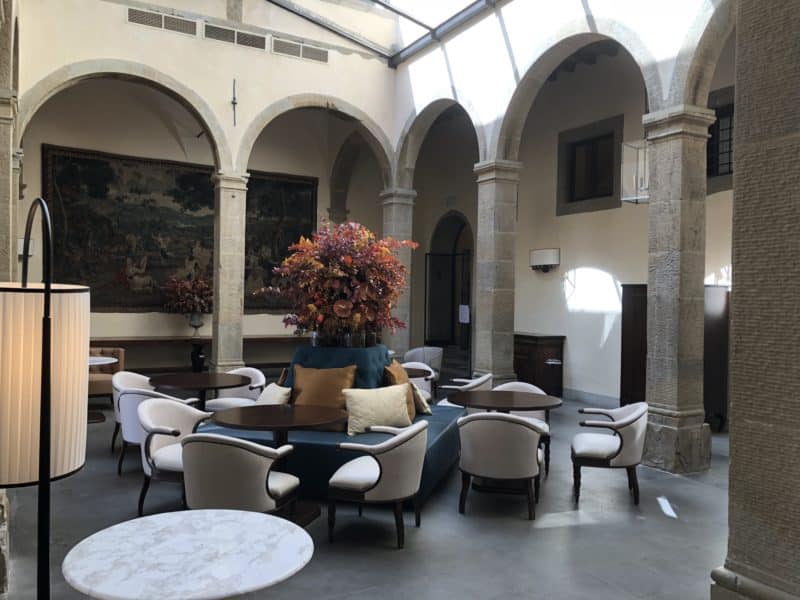 A salon area with cream and dark wood chairs clustered around a centre cushioned circular area, upon which is a tall vase full of fresh flowers. The room is filled with white stone and arches.
