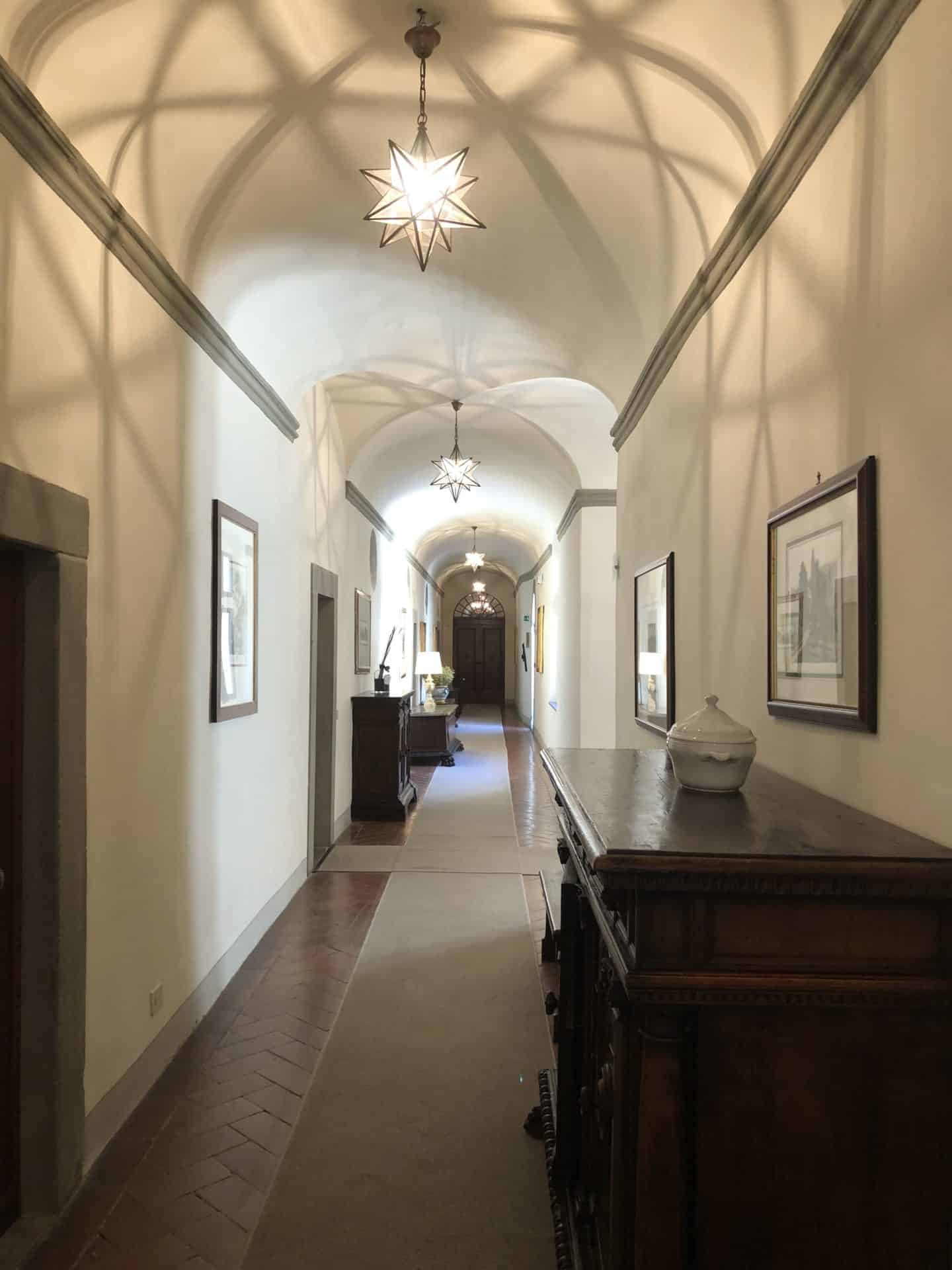 Stunning luxury hotel in Fiesole: valuted corridors of the main convent house on the first floor. There are brown dark wood cupboards on the side with photos as well as doors which lead off to the bedrooms.