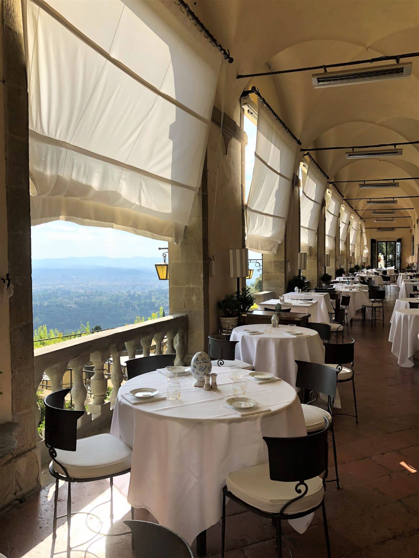 La Loggia restaurant with circular tables in a row all with white table cloths and crockery on table. There are Roman blinds on the open air Loggia arches with views looking out to the garden and Florence.