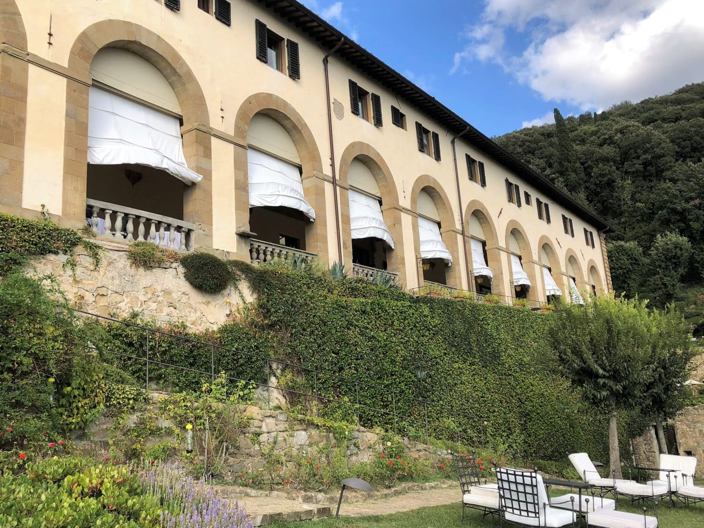 Belmond Villa San Michele: A stunning luxury hotel in Fiesole. A side view of the hotels AL Loggia restaurant with sunbeds and gardens in front of the building. The skies are blue against the beige and stone colout of the building. 