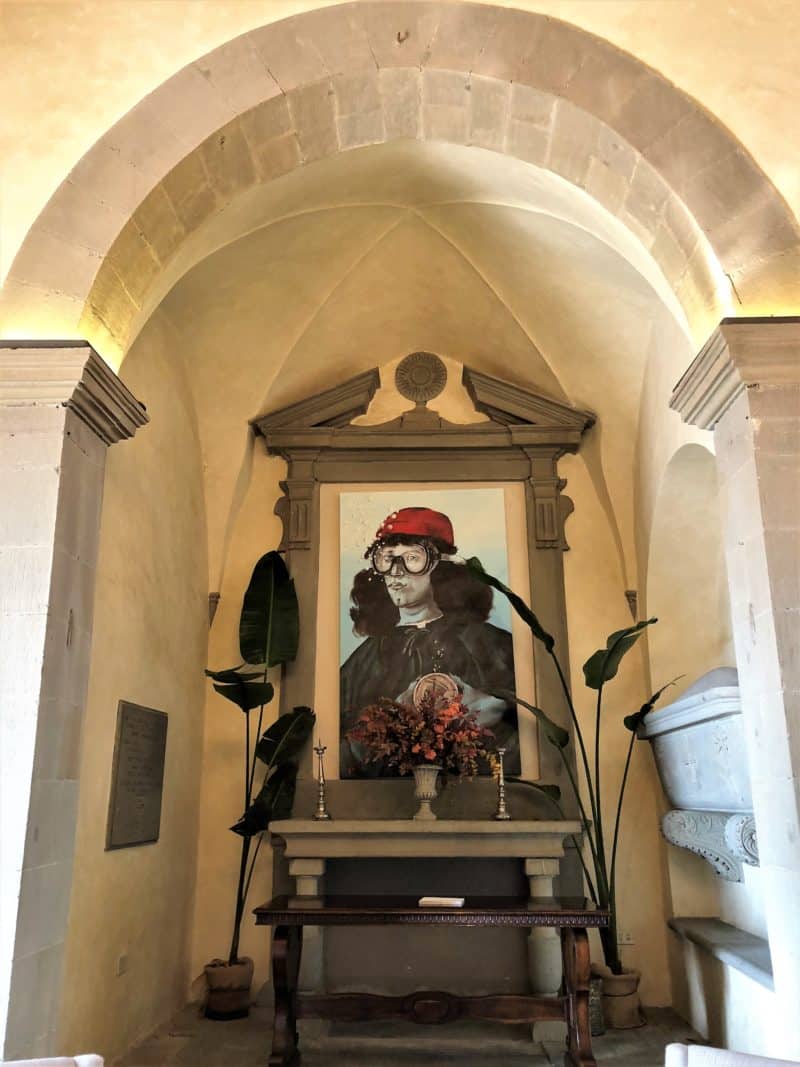 A large poece of framed wall art with a man wearing a red cap and aviator goggles. The painting rests on a table with plants at either side and an archway recess.