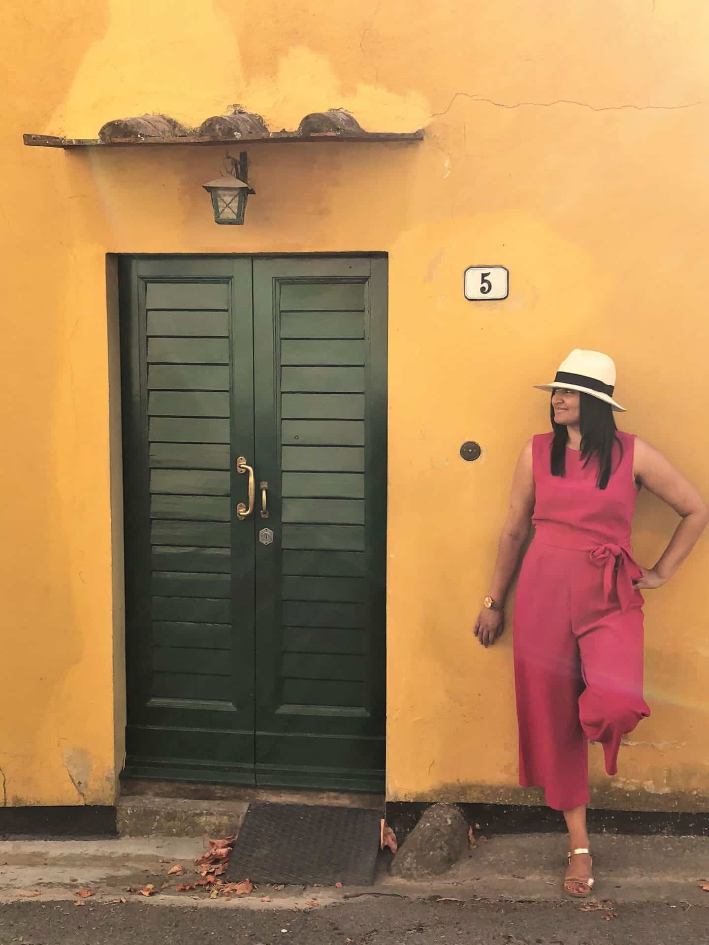 Bejal wearing a cerise pink jumpsuit and white fedora standing against a bright yellow house facade with a bottle green slatted door to her right.