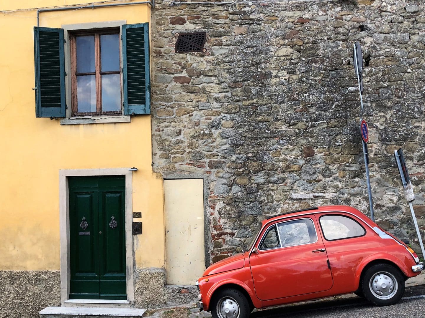 A red Fiat 500 parked at a slant against a stone wall in the village of Fiesole. There is a yellow walled house to the left which has a green door and shutters