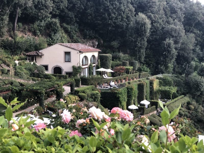 Private villa on the hotel grounds surrounded by pink and white flowers and foliage. The villa has a white/beige exterior with a terracota roof and dark brown window frames.