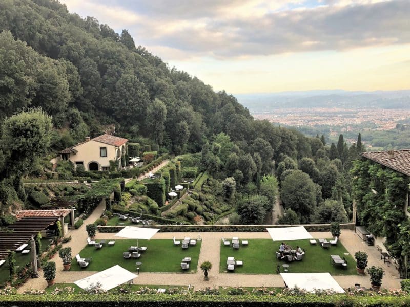 A stunning luxury hotel in Fiesole: The view sof teh hotel Loggia outdoor bar from the top tier garden. There are views of the private villa, bar area with rattan chairs , cushions and tables as well the view of Florence in the distance amongst the sea of green tress.