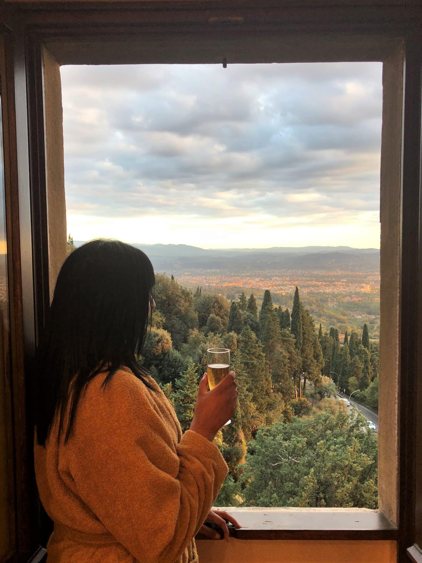 Stunning Luxury hotel in Fiesole: Bejal wearing a dressing gown and holding a glass of prosecco  looking out of bedroom window towards re sunsetting over the Fiesole hills with Florence in the distance shadowed by tall green trees.