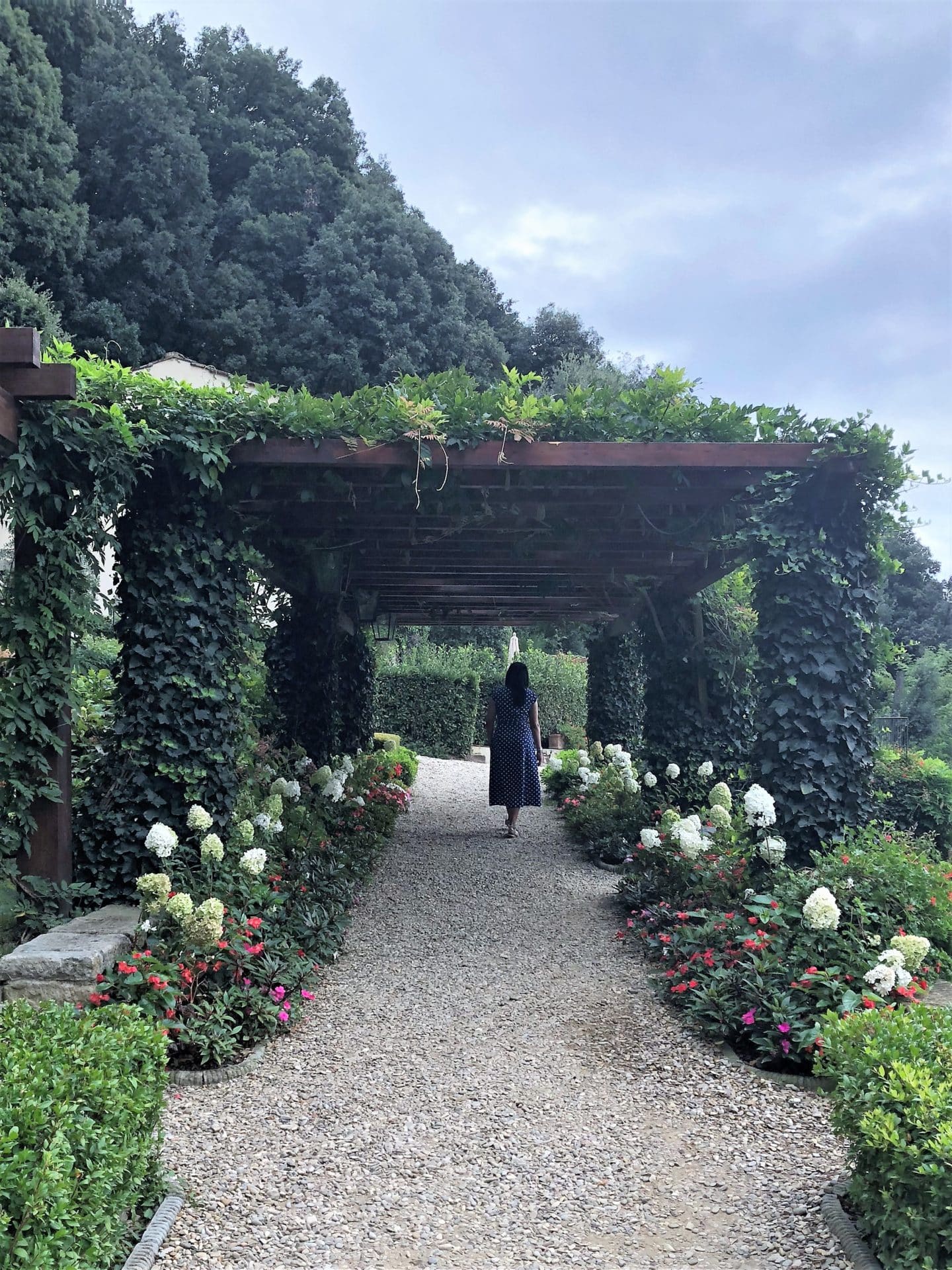 Belmond Villa San Michele: Bejal in a bright blue dress with white spots walking through the garden trellises with white roses on either side. 