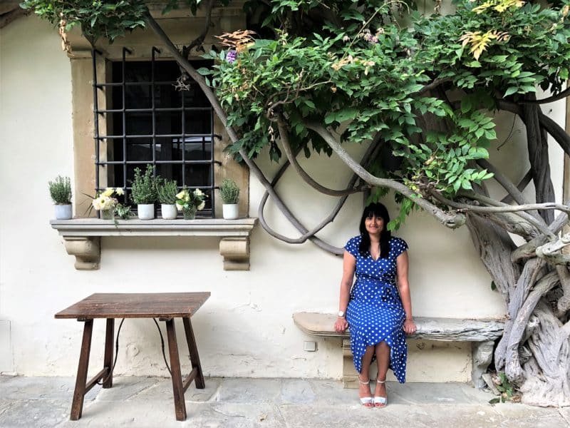 Belmond Villa San Michele: Bejal wearing a bright blue and white spot dress sitting on a bench surrounded by old vines and green leaves around. There is a window with a grid across it to Bejal's right , with some potted plants on the windowsill underneath which is a darkk wood side table.
