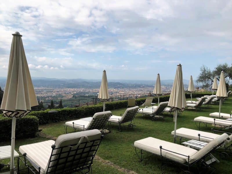 A stunning lucury hotel in Fiesole: cream cushioned sun loungers with umbrellas on the top tier of teh hotel gardens with views looking out to Florence.
