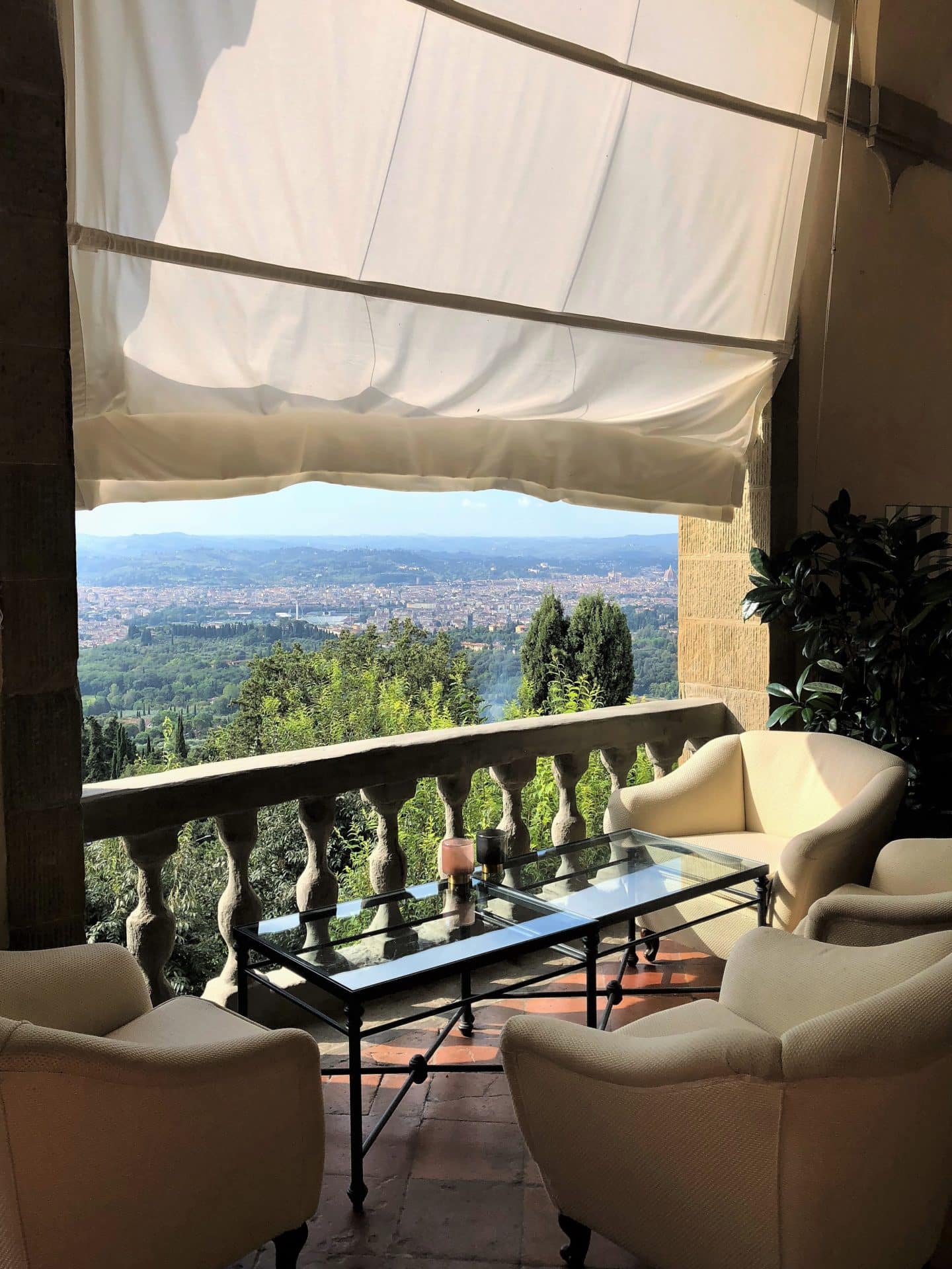 A portrait view of one of the Loggia arched windows with Roman blinds, a coffee table and 2 white armchairs with views out to the gardesn and Florenc ein the background.