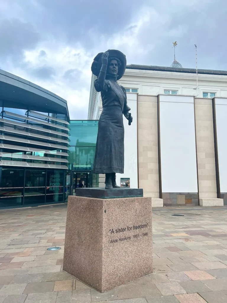 Alice Hawkins Statue at Leicester Market with the market place in the background, Leicester. The iron statue is on top of a marble square block which reads, A sister for freedom, Alice Hawkins, 1863-1946