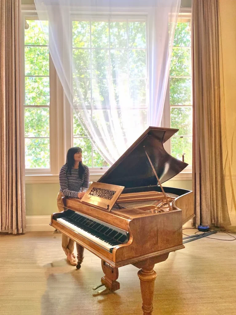 Bejal at Piano in The City Rooms, Leicester. The piano is a grand piano and Bejal is leaning agains the piano with a large window behind letting in lots of light
