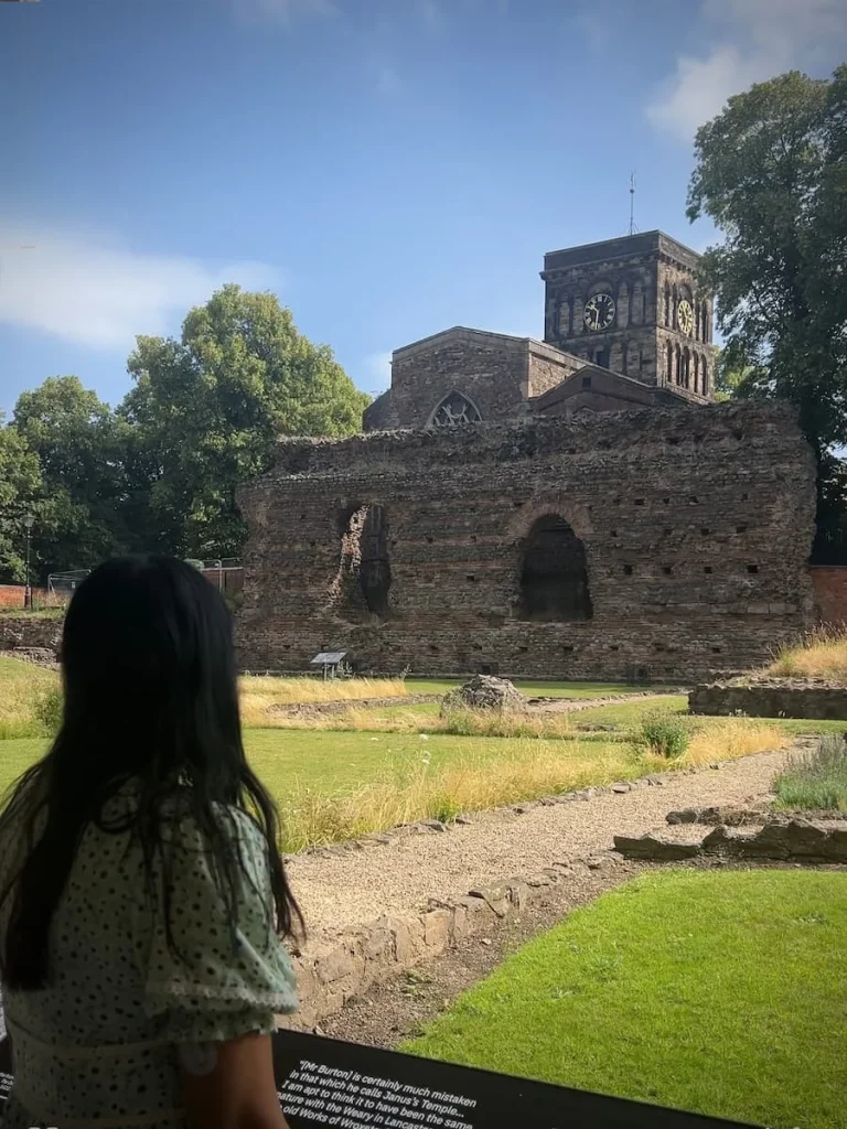 Bejal standing in front of Jewry Wall, Leicester on a sunny day with the grass and wall remainders on the ground. Bejal has her back to the camera