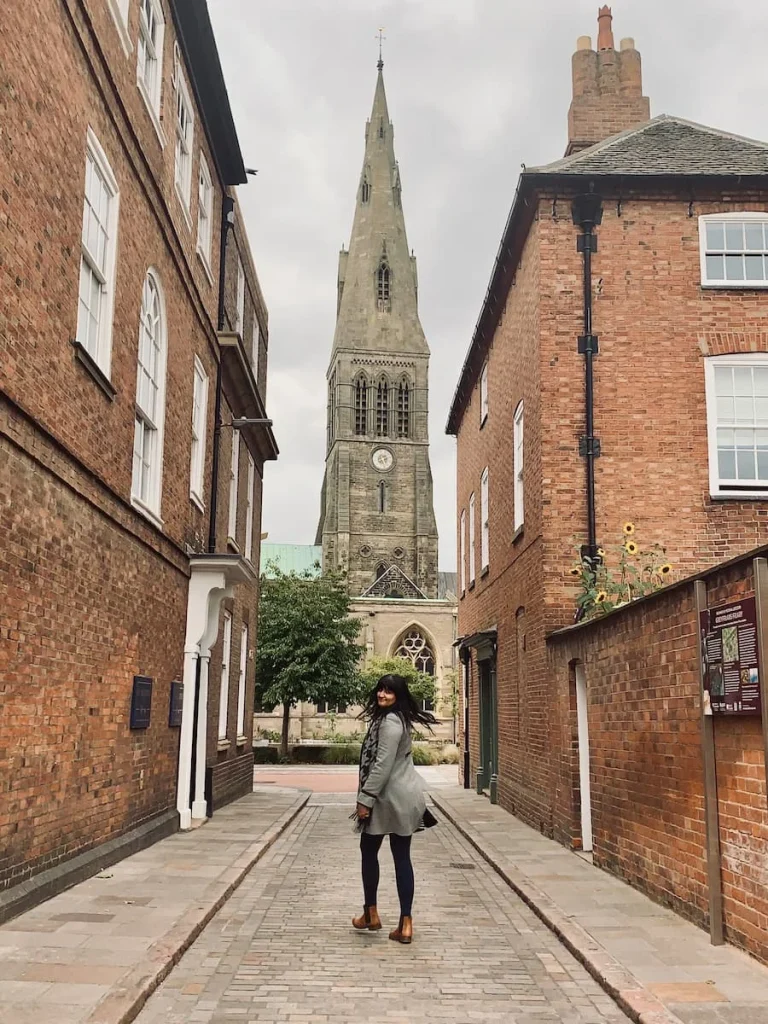Bejal walking down New Street with the cathedral spire right in front of her on a cobbled stoned street. There are red brick buildings at either side. Bejal is wearing a grey coat, black tights and tan Chelsea boots