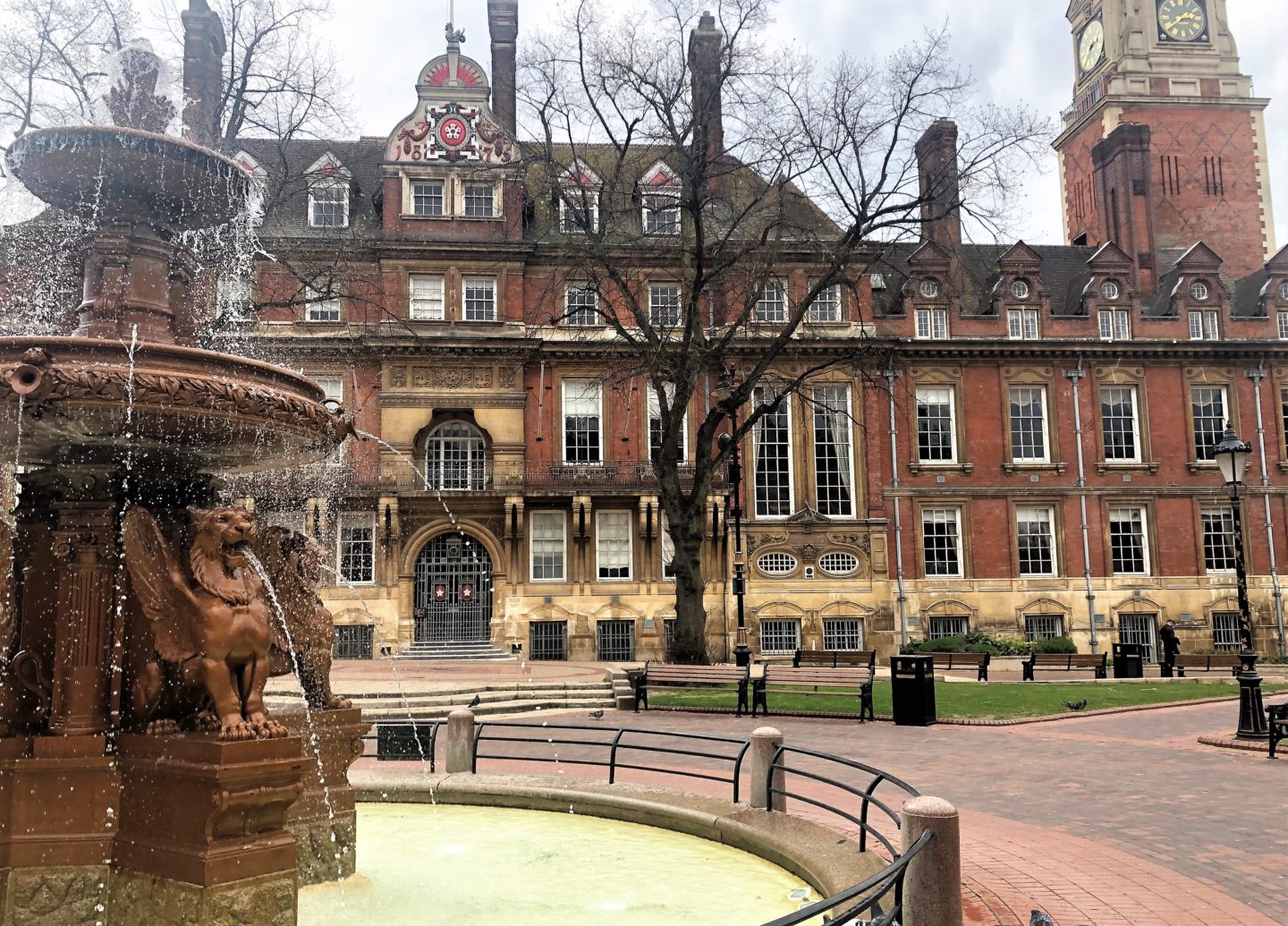 The town hall square fountain with water running from the lions mouths. The town hall square is in the background with benches and its clock tower in the background.