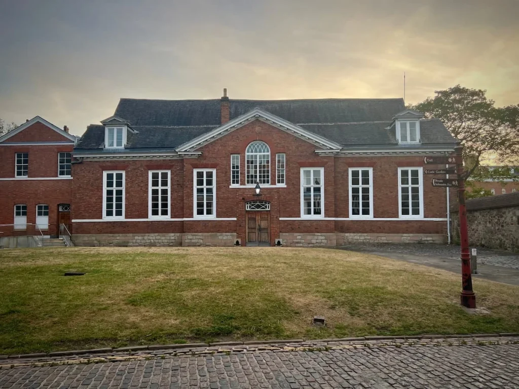 The exterior of Leicester Castle with its red brick building and modern slate roof and tall white window sills. There is a small grass mound right infront of the building.