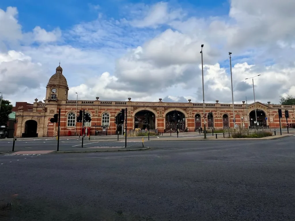 Leicester Railway Station taken across the street with the clock face dome and full length of the station in red and cream brick and stone work. The blue sky is in the background with bus stops in front of the station