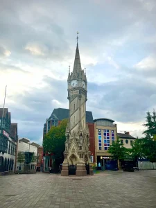 Leicester clock Tower standing tall with ints grey stone work on East Gates, Leicester. The clock is at the top of teh tower and shops and trees surround the tower on a small sqaure