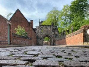 Ruperts Gatweay with cobbled-stones in front. It's possible to see St Mary de Castro tower in the far distance as well as the brick walls of Newarke Houses Museum and Trinity House Herb Garden. The sky is blue with a few clouds