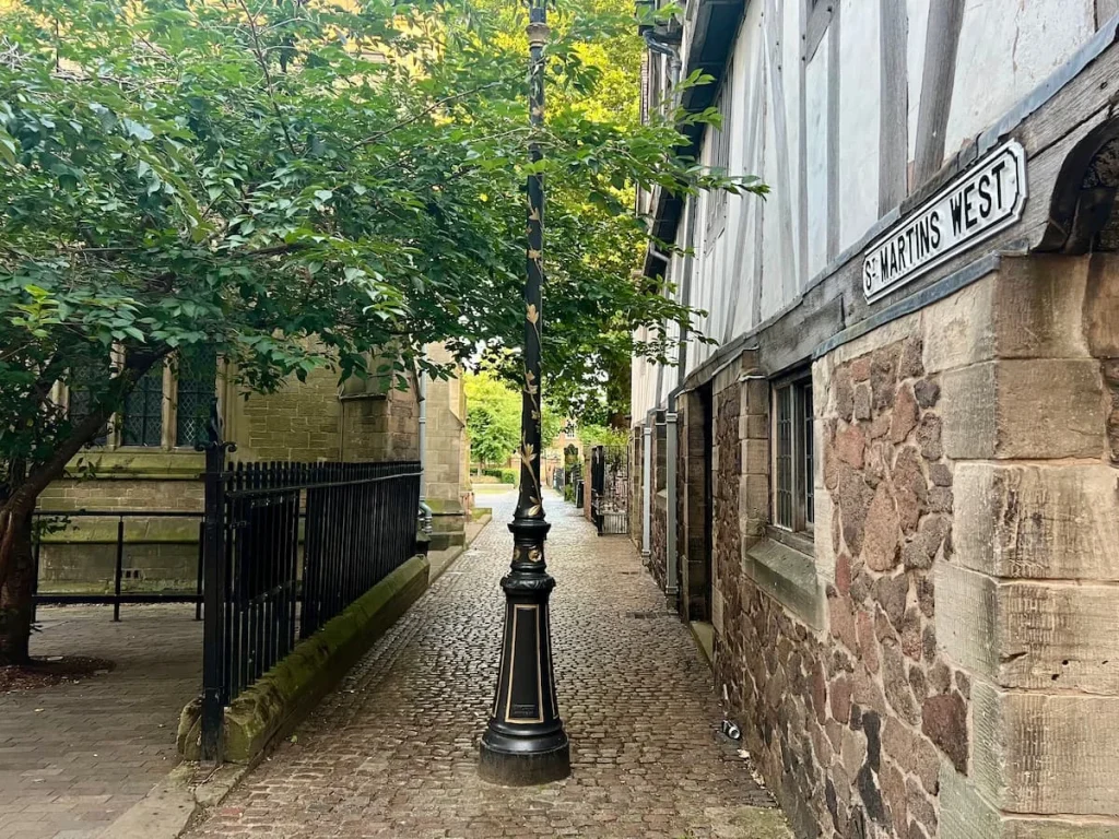 St Martin's West Lane with a Victorian lamppost, black railings adn a tree on one side with the Guildhall on the right, framing the image. The floor is greYcobble stones.