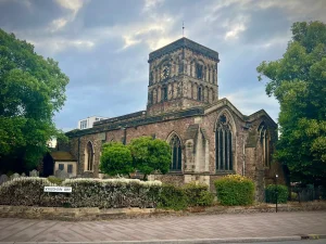 The exterior of St.Nicholas Church in Leicester with it's romanesque tower and stained glass window. and graveyard gardens. There is a road sign reading Vaughn Way on the fence outside