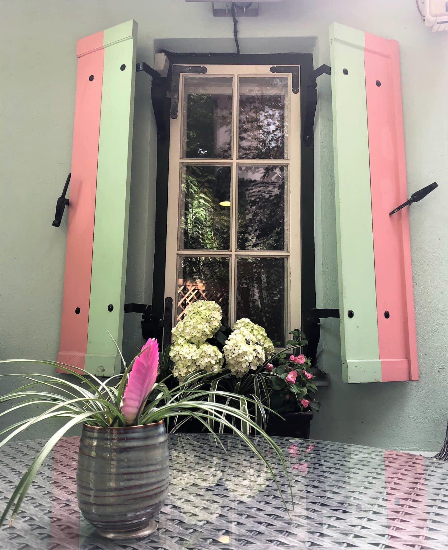 The Schlossle Hotel Tallinn: A slim windown with pale green and candy pink shutters. There are some white hydrangeas in a window box and a vase of flowers on the rattan table of the garden furniture. View is from he summer garde courtyard at the hotel.