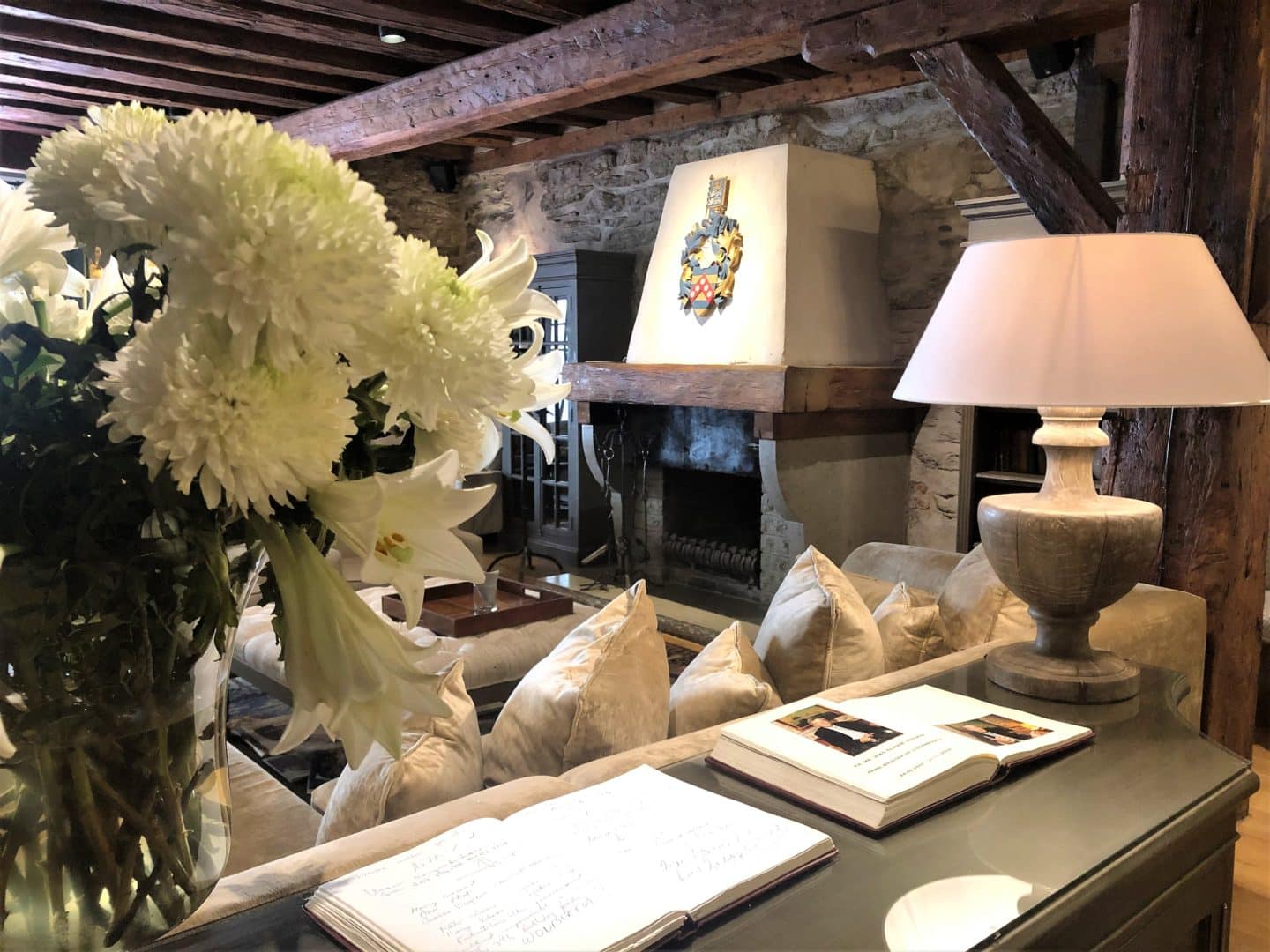 The Schlossle Hotel Tallinn: The lounge and lobby with beamed brown ceilings, a huge comfy corner sofa and stoned walls. There is a bunch of white flowers framing the photo and a white lampshade with a stone base, with the fireplace in the background