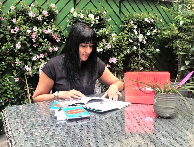 The Schlossle Hotel Tallinn: Bejal sitting in the summer garden with pink flowers behind here planning a city itinerary with a Visit Tallinn red bag