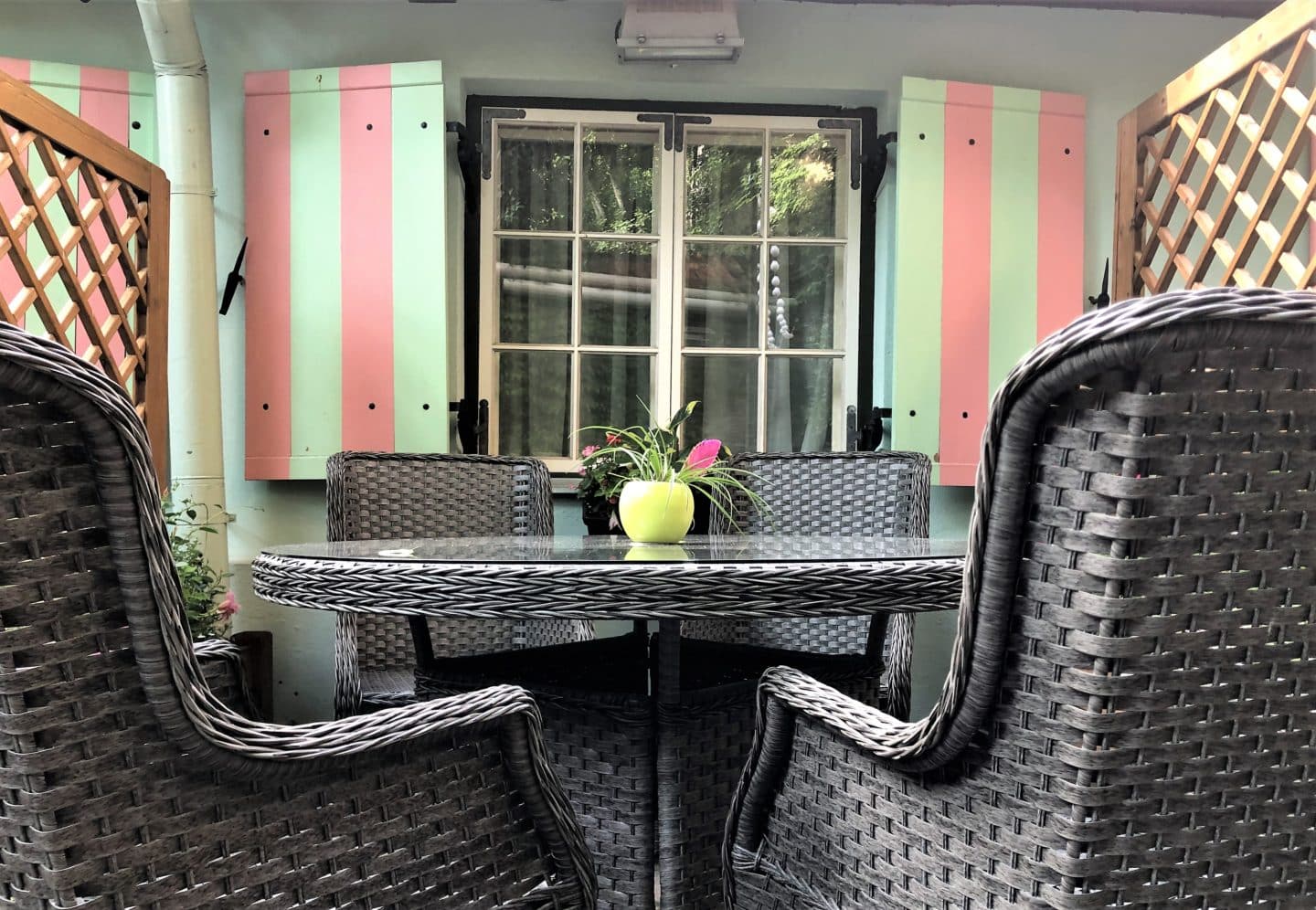 The Schlossle Hotel Tallinn: grey rattan chairs with a rattan table and a small vase with flowers in it. There is a window in the background with a pale green and pink striped wall.