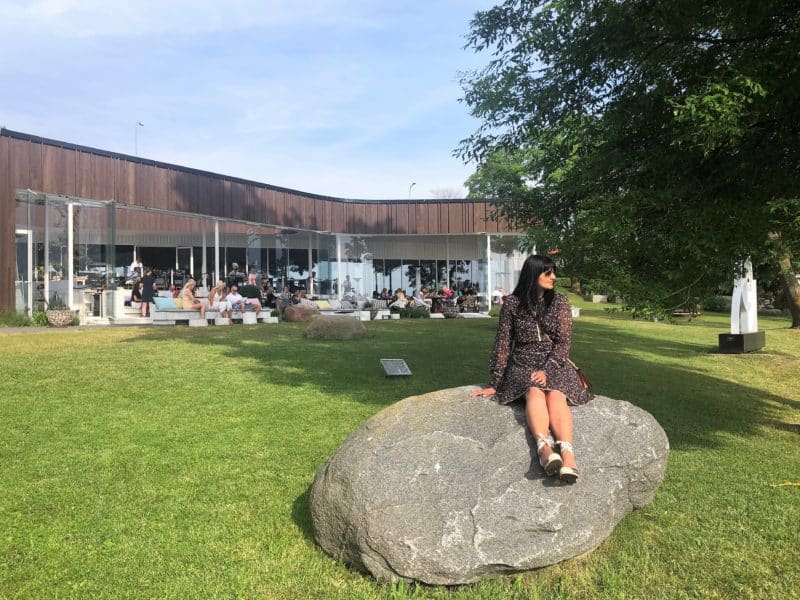 Bejal sitting on a large stone outside the back end of Noa Restorans, surrounded by trees and grass. Bejal is looking out to the waters of Tallinn Bay with other diners in the background