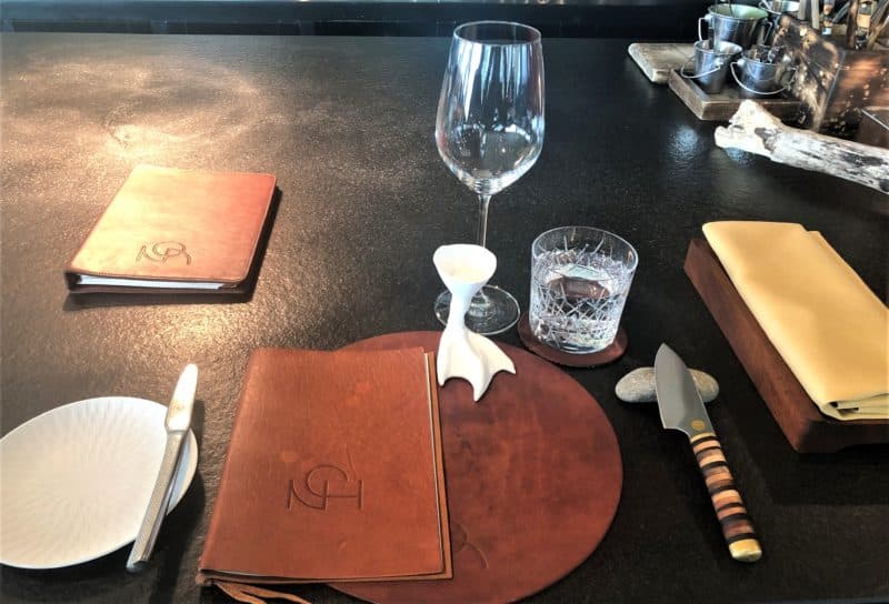 The che's counter at Noa Chef's Hall in Tallinn, dressed for dinner with tan leather brown menus and matching place mats with water glasses and a wine glass. There is a yellow cloth napkin and silver cutlery on the table. 